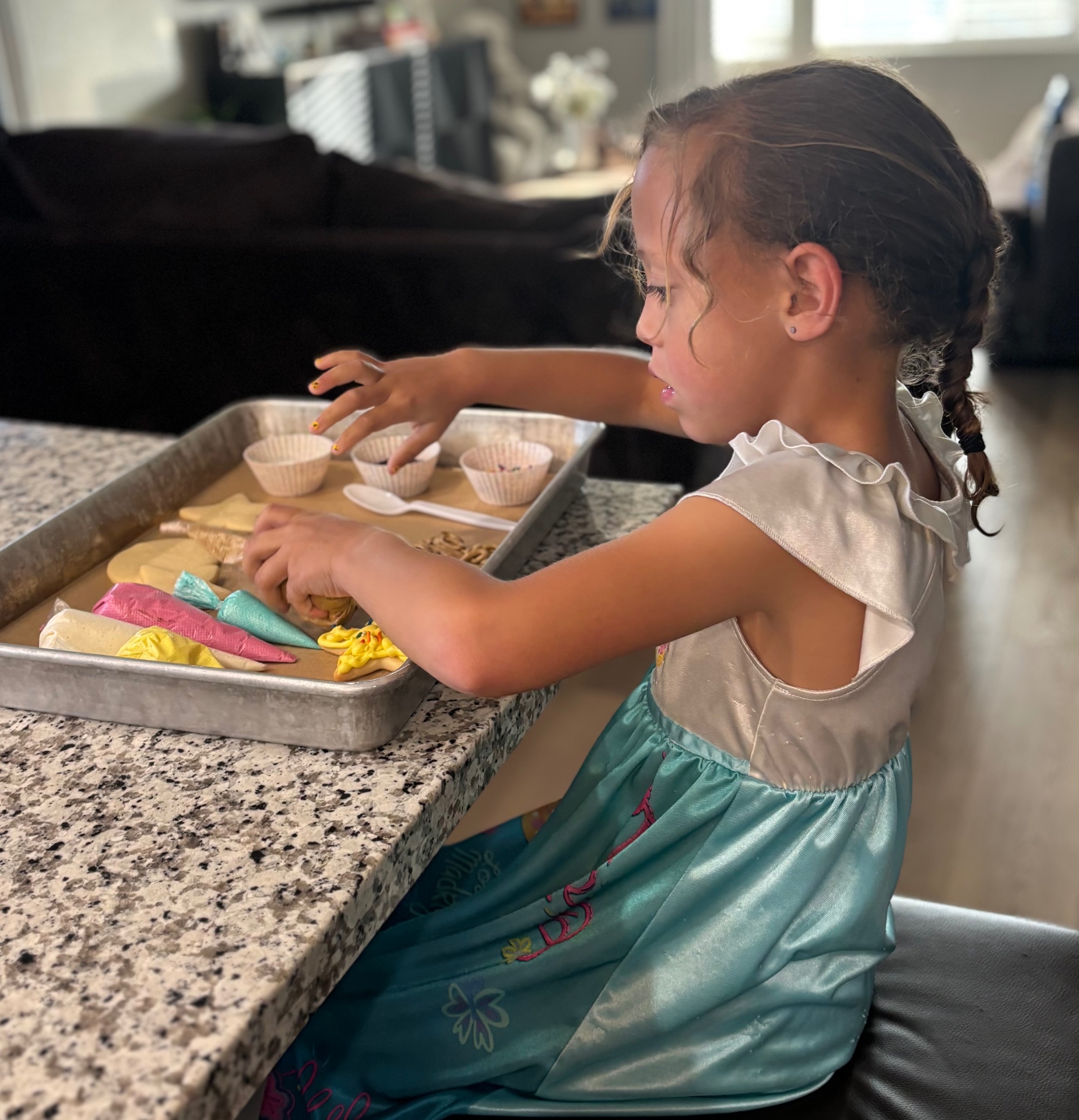 A young girl in a teal dress decorating sugar cookies at the kitchen counter, surrounded by frosting bags and sprinkle cups